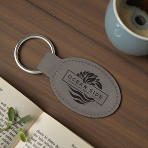 Gray leather keychain with 'Ocean Side Commercial Realty Group' logo on a wooden surface with a book and coffee cup.