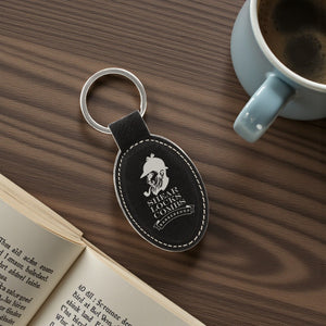 Black leather keychain with logo on a wooden surface next to an open book and a cup of coffee.