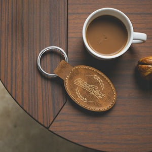 Brown leather keychain with 'Oktoberfest' engraving next to a cup of coffee on a wooden table.