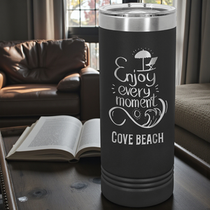Black tumbler with 'Enjoy every moment at Cove Beach' text on a table with a book and sofa in the background.