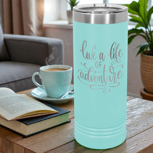 Teal tumbler with 'live a life of adventure' text on a table with a cup of coffee and a book.