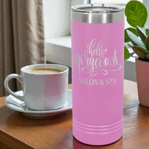 Pink tumbler with 'hello gorgeous' and 'SALON & SPA' text next to a white mug with coffee on a wooden table.