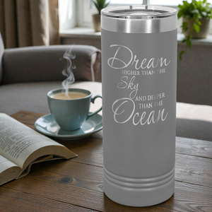 Gray tumbler with motivational quote next to a steaming cup of coffee on a table.