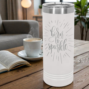 White tumbler with 'born to sparkle' text on a wooden table with a cup of coffee and book.