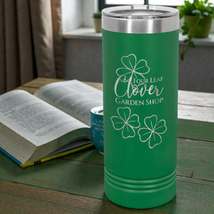Green tumbler with 'Four Leaf Clover Garden Shop' design on a wooden table with an open book and plant in the background.