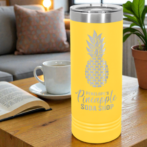 Yellow tumbler with pineapple design and text on a table with a cup and book in a cozy living room.