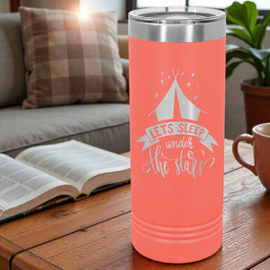 Coral tumbler with a camping design and text on a wooden table with a couch and book in the background.